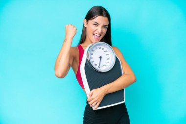 Mavi arka planda, tartı makinesiyle izole edilmiş beyaz bir kadın zafer jesti yapıyor.