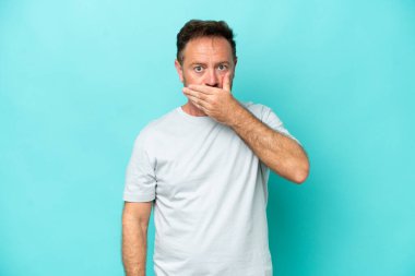 Orta yaşlı, beyaz bir adam, mavi arka planda izole edilmiş, ağzı elle kapatılmış.
