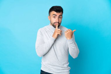 İzole edilmiş mavi arka planda beyaz bir adam. Yan tarafı işaret ediyor ve sessizlik hareketi yapıyor.