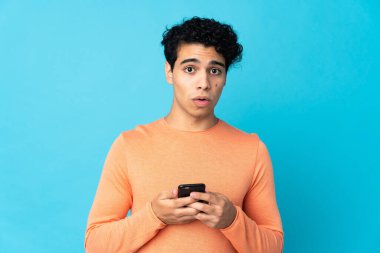 Venezuelalı adam mavi arka planda izole edilmiş kameraya bakarken şaşırmış bir ifadeyle cep telefonunu kullanıyor.