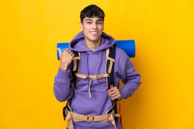 İzole edilmiş sarı arka planı olan büyük bir sırt çantasıyla genç dağcı bir ürünü sunmak için kenarı işaret ediyor.