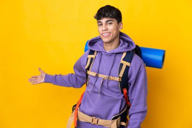İzole edilmiş sarı arka plan üzerinde büyük bir sırt çantası olan genç dağcı, davet etmek için ellerini yana uzatıyor.