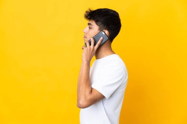 İzole edilmiş sarı arka planda genç bir adam cep telefonuyla sohbet ediyor.