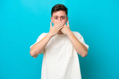 Genç ve yakışıklı Brezilyalı bir adam mavi arka planda izole edilmiş, ağzı elleriyle kapatıyor.