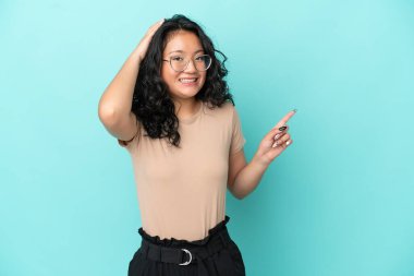 Asyalı genç bir kadın mavi arka planda izole edilmiş. Şaşırmış ve işaret ediyor.