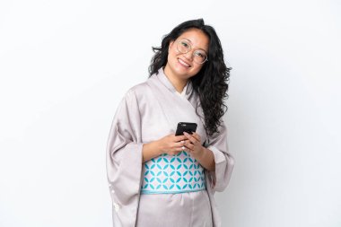 Kimono giyen genç Asyalı kadın beyaz arka planda izole edilmiş cep telefonuyla bir mesaj gönderiyor.