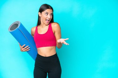 Genç beyaz kadın yoga derslerine giderken elinde mavi arka planda bir minder tutarken sürpriz bir ifadeyle yan tarafa bakıyor.
