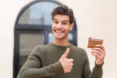 Açık havada cüzdan tutan genç beyaz bir adam. Başparmakları havada. Çünkü iyi bir şey oldu.