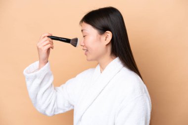 Genç Çinli kadın bej arka planda izole edilmiş elinde makyaj fırçası ve Whit mutlu yüz ifadesi var.