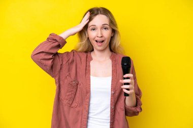 Sarı arka planda, sürpriz bir ifadeyle izole edilmiş bir mikrofonu kaldıran beyaz bir kadın.