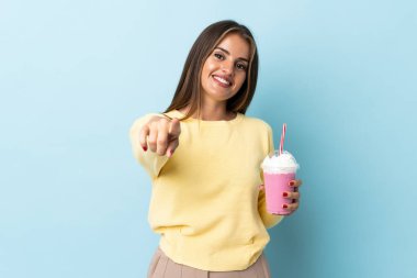 Mavi arka planda izole edilmiş çilekli milkshake ile genç Uruguaylı kadın mutlu bir ifadeyle öne bakıyor.