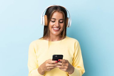 Genç Uruguaylı kadın mavi arka planda izole edilmiş müzik dinliyor ve cep telefonu arıyor.