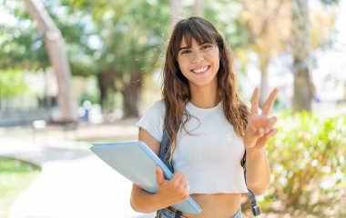 Açık havada gülümseyen ve zafer işareti gösteren genç bir bayan öğrenci.