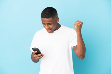 Genç Latin adam mavi arka planda cep telefonu kullanarak ve zafer jesti yaparak izole edildi.
