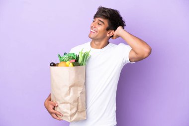 Elinde alışveriş çantası tutan genç bir adam mor arka planda izole edilmiş, çok gülümsüyor.