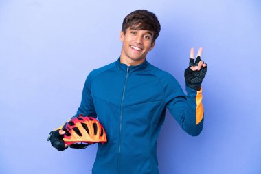 Mor arka planda izole edilmiş genç beyaz adam gülümsüyor ve zafer işareti gösteriyor.