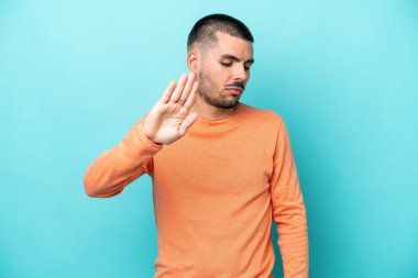 Mavi arka planda izole edilmiş genç beyaz adam dur hareketi yapıyor ve hayal kırıklığına uğruyor.