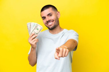 Kafkasyalı genç bir adam, sarı arka planda izole edilmiş bir sürü para alıyor mutlu bir ifadeyle ön tarafı gösteriyor.