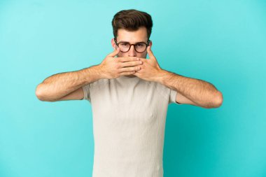 Beyaz tenli yakışıklı adam mavi arka planda izole edilmiş, ağzı elleriyle kapatıyor.
