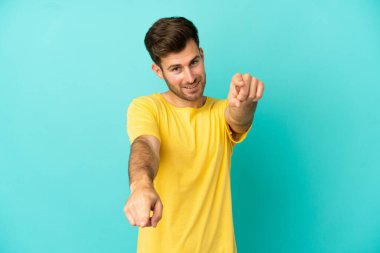 Genç beyaz yakışıklı adam mavi arka planda izole edilmiş mutlu bir ifadeyle ön tarafı işaret ediyor.