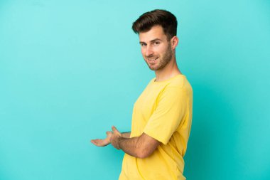 Beyaz tenli yakışıklı adam mavi arka planda izole edilmiş. Davet etmek için ellerini yana uzatıyor.