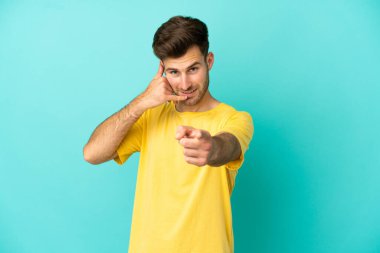 Beyaz tenli yakışıklı bir adam mavi arka planda izole edilmiş telefon hareketlerini yapıyor ve ön tarafı işaret ediyor.