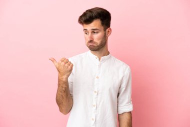 Pembe arka planda izole edilmiş genç beyaz yakışıklı adam mutsuz ve bir tarafı işaret ediyor.