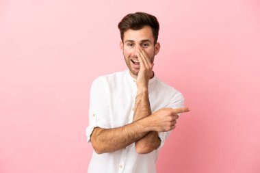 Pembe arka planda soyutlanmış beyaz tenli yakışıklı bir adam bir ürün sunmak ve bir şeyler fısıldamak için kenarı işaret ediyor.