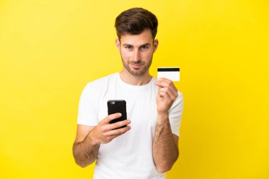 Genç beyaz yakışıklı adam sarı arka planda izole edilmiş cep telefonuyla kredi kartıyla alışveriş yapıyor.
