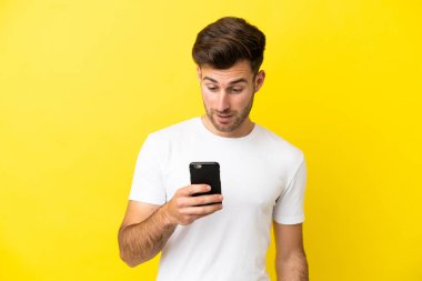 Sarı arka planda izole edilmiş genç beyaz adam cep telefonuyla mesaj ya da e-posta gönderiyor.