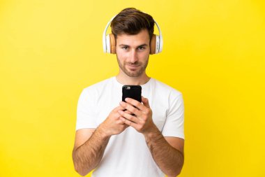 Beyaz tenli yakışıklı adam sarı arka planda izole edilmiş. Cep telefonuyla müzik dinliyor ve ön tarafa bakıyor.