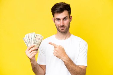 Young caucasian man taking a lot of money isolated on yellow background pointing to the side to present a product