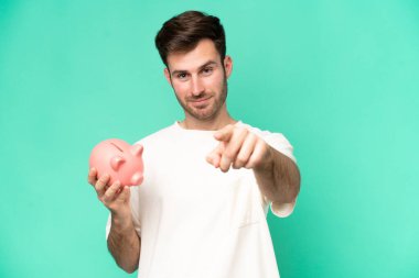 Kumbarayı elinde tutan genç beyaz adam yeşil arka planda izole edilmiş. Kendine güvenen bir ifadeyle sana parmak basıyor.