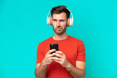 Kafkasyalı genç adam mavi arka planda izole edilmiş. Cep telefonuyla müzik dinliyor ve düşünüyor.