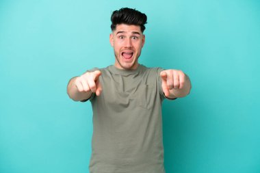 Mavi arka planda izole edilmiş genç beyaz adam şaşırmış ve ön tarafı işaret ediyor.