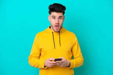 Mavi arka planda izole edilmiş genç beyaz adam şaşırdı ve bir mesaj gönderiyor.
