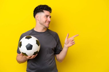 Yakışıklı genç futbolcu, sarı arka planda izole edilmiş, parmağıyla kenarı işaret ediyor.