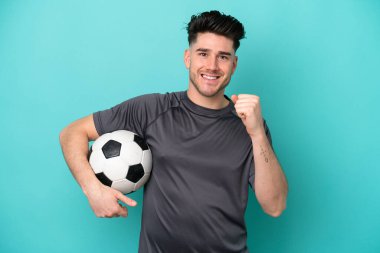 Genç beyaz adam mavi arka planda izole edilmiş bir şekilde futbol topuyla zaferi kutluyor.