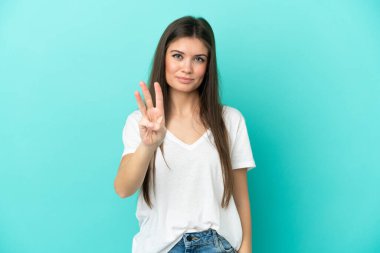 Mavi arka planda izole edilmiş genç beyaz kadın mutlu ve parmaklarıyla üçe kadar sayıyor.