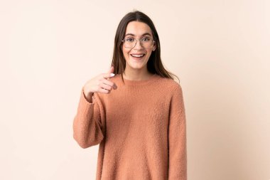 İzole edilmiş bej arka planı olan genç bir kadın şaşırmış ve ön tarafı işaret ediyor.