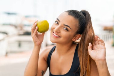 Açık havada genç ve güzel bir kadın elinde mutlu bir ifadeyle bir elma tutuyor.