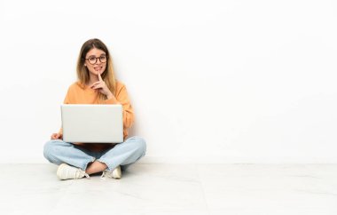 Dizüstü bilgisayarı olan genç bir kadın yerde sessizce oturuyor ve parmağını ağzına sokuyor.