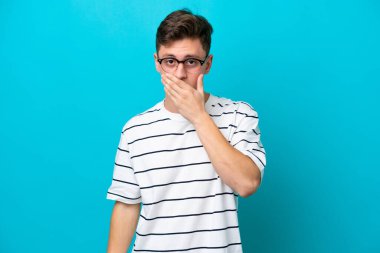 Genç ve yakışıklı Brezilyalı bir adam mavi arka planda izole edilmiş. Ağzını eliyle kapatıyor.