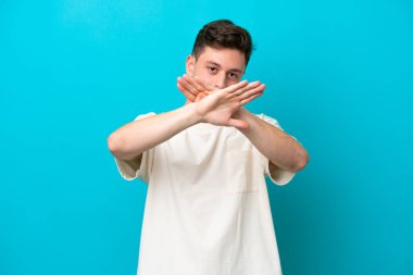 Genç, yakışıklı Brezilyalı bir adam, mavi arka planda izole edilmiş bir şekilde, bir eylemi durdurmak için eliyle jest yapıyor.