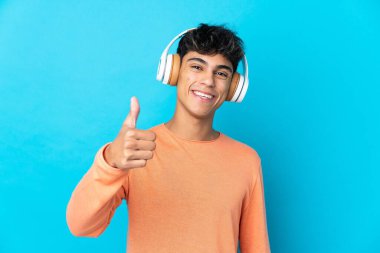 Genç adam izole edilmiş mavi arka planda müzik dinliyor ve başparmağını kaldırıyor.