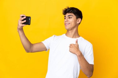 İzole edilmiş sarı arka planı olan genç bir adam cep telefonuyla selfie çekiyor.
