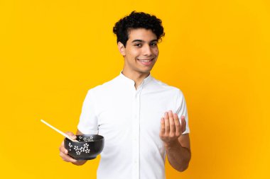 Genç Venezuelalı bir adam sarı arka planda izole edilmiş ve bizimle gelmesi için davet ediliyor. Yemek çubuğuyla bir kase erişte tutarken geldiğin için çok mutluyum.