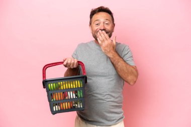 Elinde alışveriş sepeti olan orta yaşlı beyaz bir adam pembe arka planda izole edilmiş mutlu ve ağzı eliyle gülümsüyor.