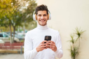 Genç beyaz bir adam açık havada müzik dinliyor cep telefonu ve ön tarafa bakıyor.