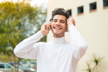 Genç beyaz adam açık havada müzik dinliyor ve şarkı söylüyor.